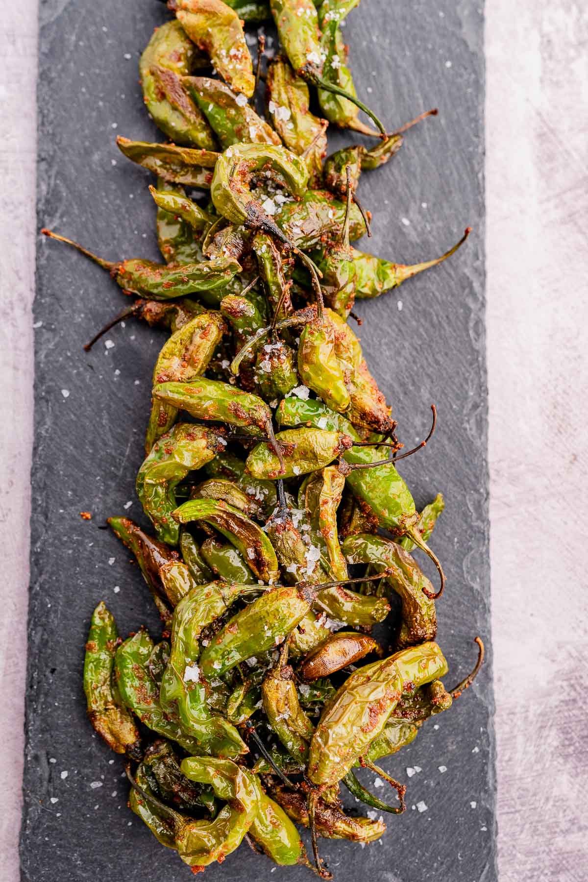 A pile of air fryer shishito peppers, roasted to perfection and sprinkled with coarse salt, rests enticingly on a rectangular slate platter.