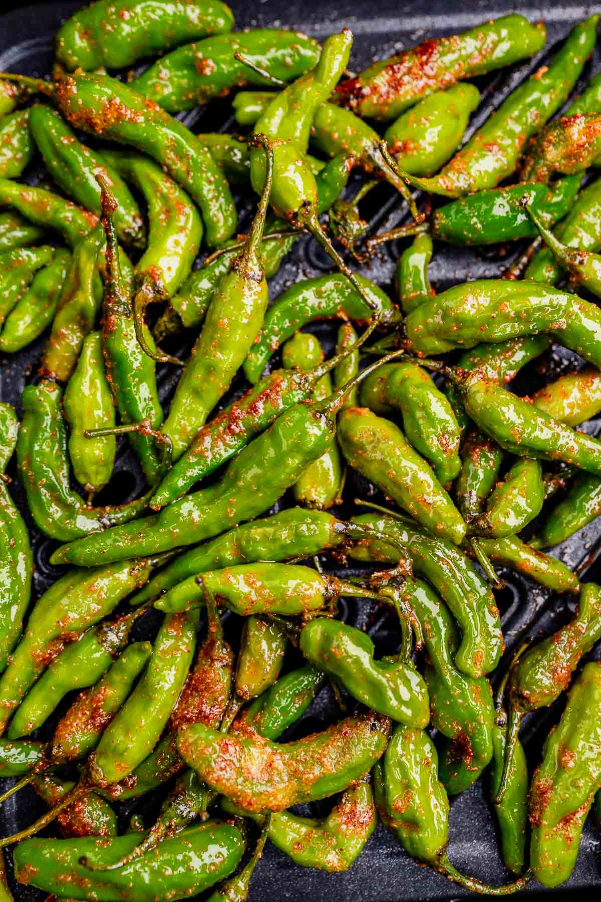 A pile of glossy, air fryer shishito peppers, seasoned with red spices, is displayed on a dark surface.