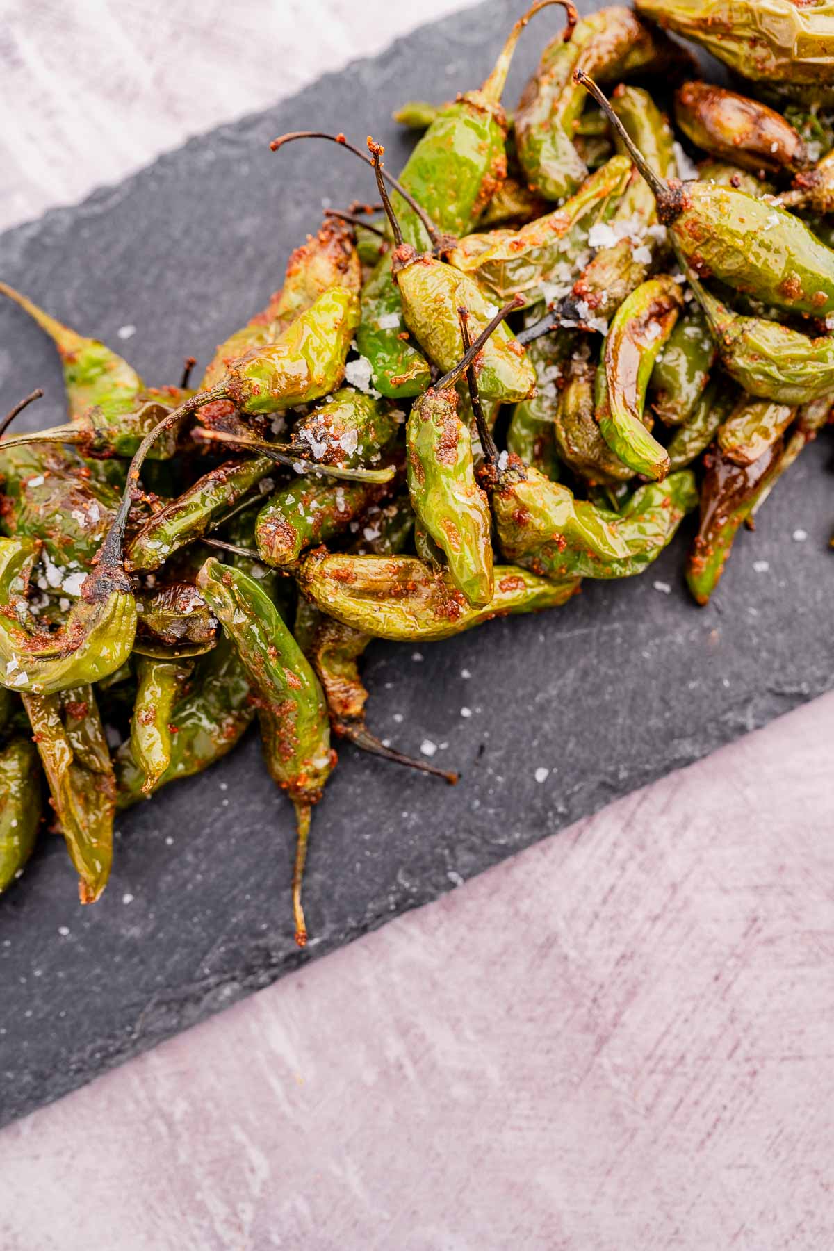 Air fryer-roasted shishito peppers garnished with coarse salt on a rectangular slate platter.