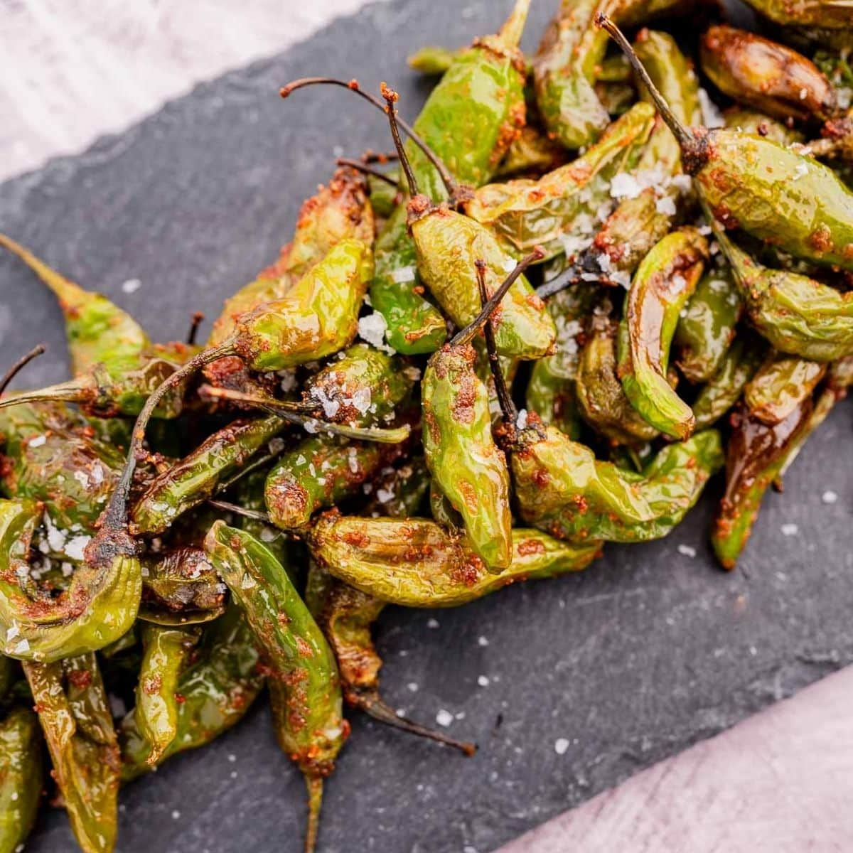 A pile of roasted green peppers sprinkled with coarse salt on a dark slate surface.