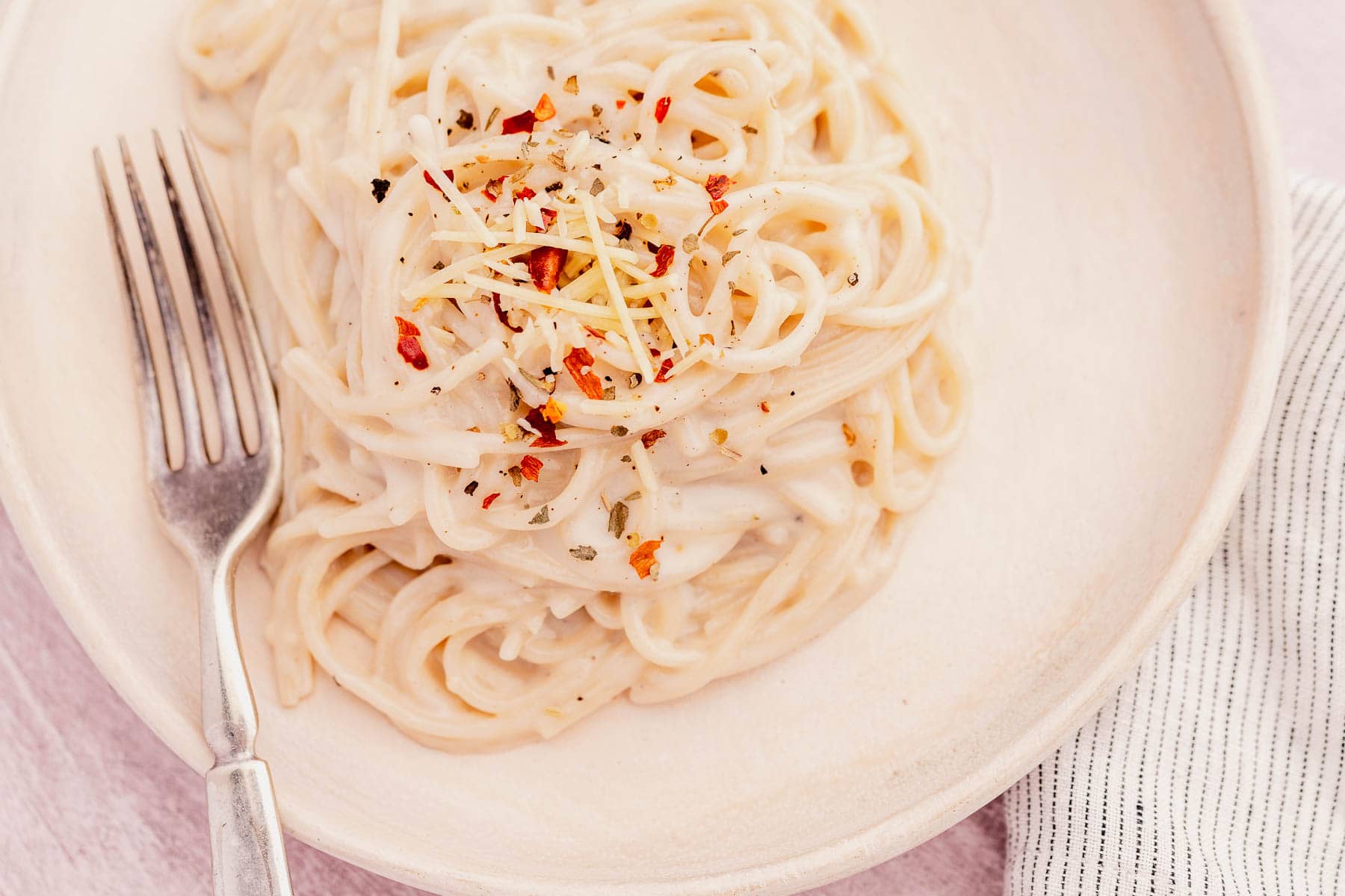 A plate of creamy garlic parmesan pasta garnished with grated cheese, chili flakes, and herbs sits invitingly next to a fork.