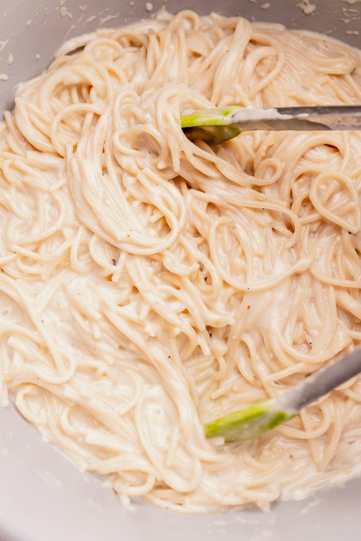 A bowl of spaghetti mixed with creamy white sauce, stirred with metal tongs.