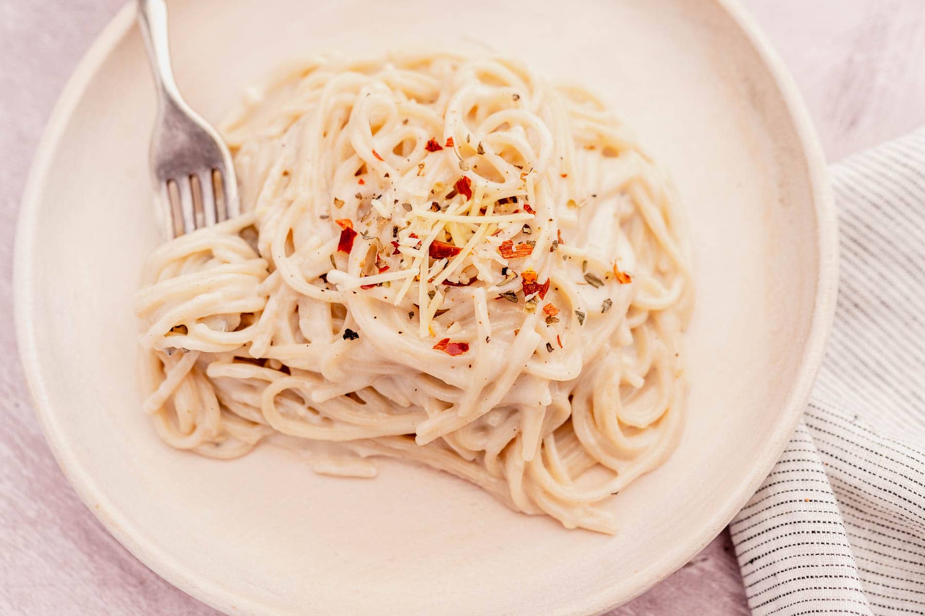 A plate of creamy garlic parmesan pasta topped with shredded cheese and red pepper flakes, served with a fork. A striped cloth is partially visible on the side.