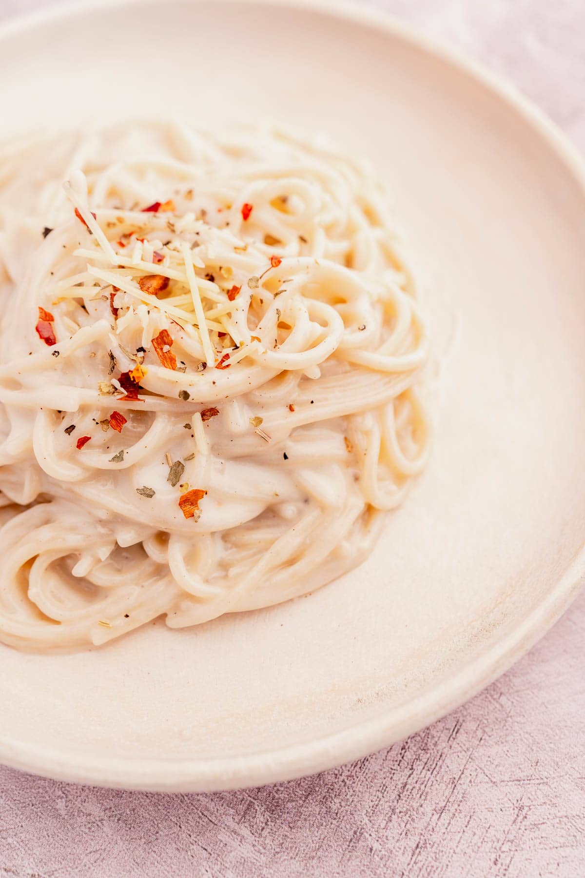 A plate of creamy pasta topped with shredded cheese and red pepper flakes on a light-colored dish.