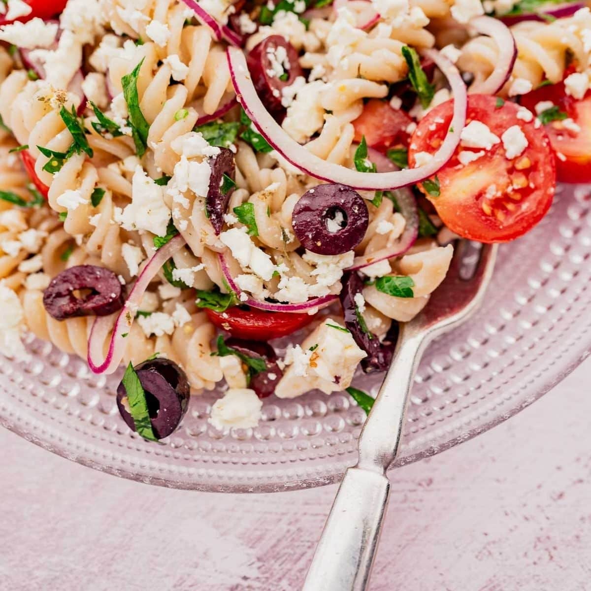 A plate of pasta salad with rotini, cherry tomatoes, feta cheese, black olives, red onion, and parsley, served with a fork.