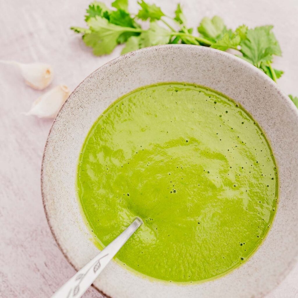 A bowl of creamy green soup with a spoon, garnished with cilantro leaves. Garlic cloves are placed nearby on a light surface.