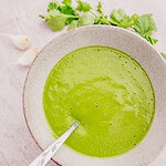 A bowl of creamy green soup with a spoon, garnished with cilantro leaves. Garlic cloves are placed nearby on a light surface.