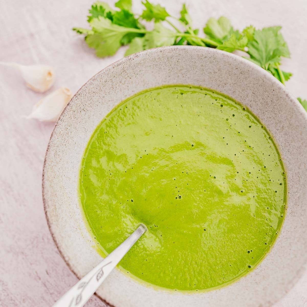 A bowl of creamy green soup with a spoon, garnished with cilantro leaves. Garlic cloves are placed nearby on a light surface.