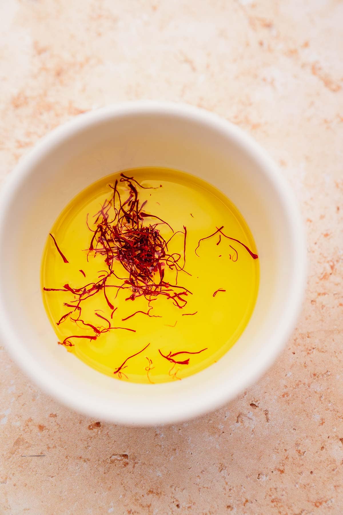 A white bowl contains a yellow saffron-infused liquid, reminiscent of dill rice's fragrant allure, with red saffron strands floating on top, placed on a textured beige surface.