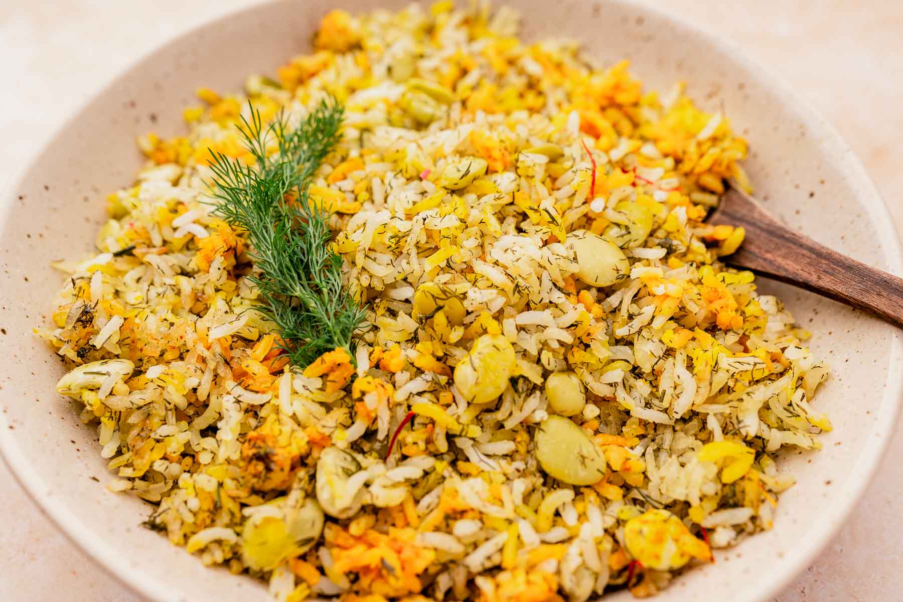 A bowl of dill rice infused with saffron and lima beans, garnished with a sprig of fresh dill, and served with a wooden spoon.