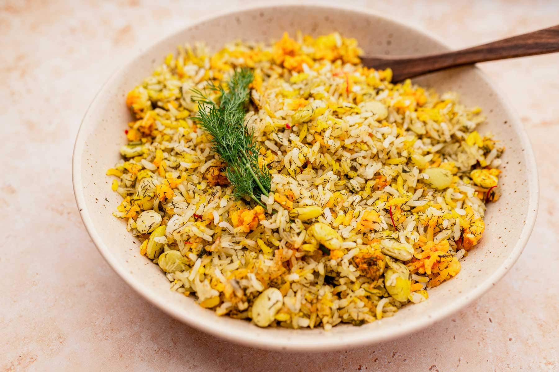 A bowl of dill rice infused with edamame, herbs, and saffron is elegantly garnished with a sprig of fresh dill on a light-colored surface.