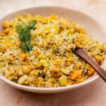 A bowl of seasoned rice mixed with vegetables, garnished with fresh dill, and served with a wooden spoon on a beige surface.