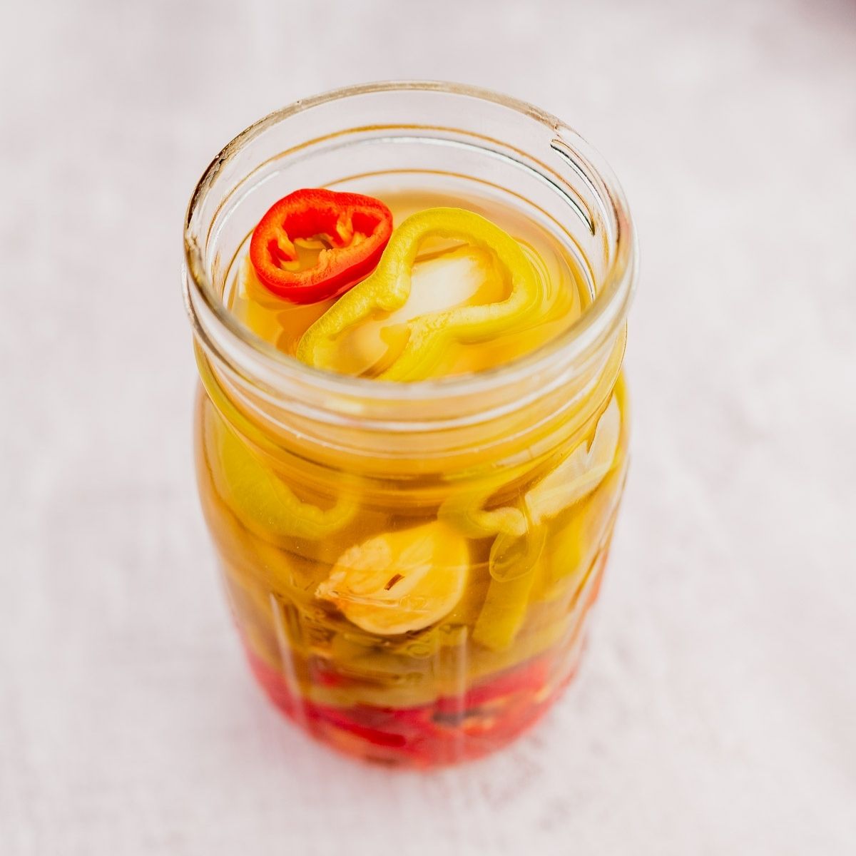 A glass jar filled with slices of pickled peppers in various colors, including red and yellow, on a light surface.