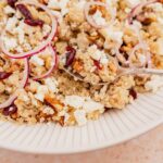 A close-up of a quinoa salad with cranberries, walnuts, red onion slices, and crumbled feta on a white plate. A spoon is scooping the salad.