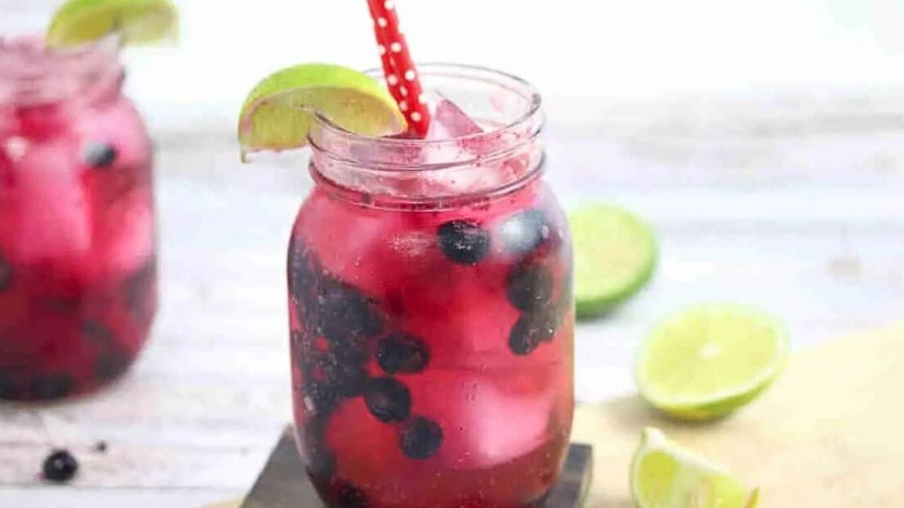 Blueberry mocktail in a mason jar with limes on the side of the jar and red straws with the jar on a wooden coater and cutting board and ingredients around it.