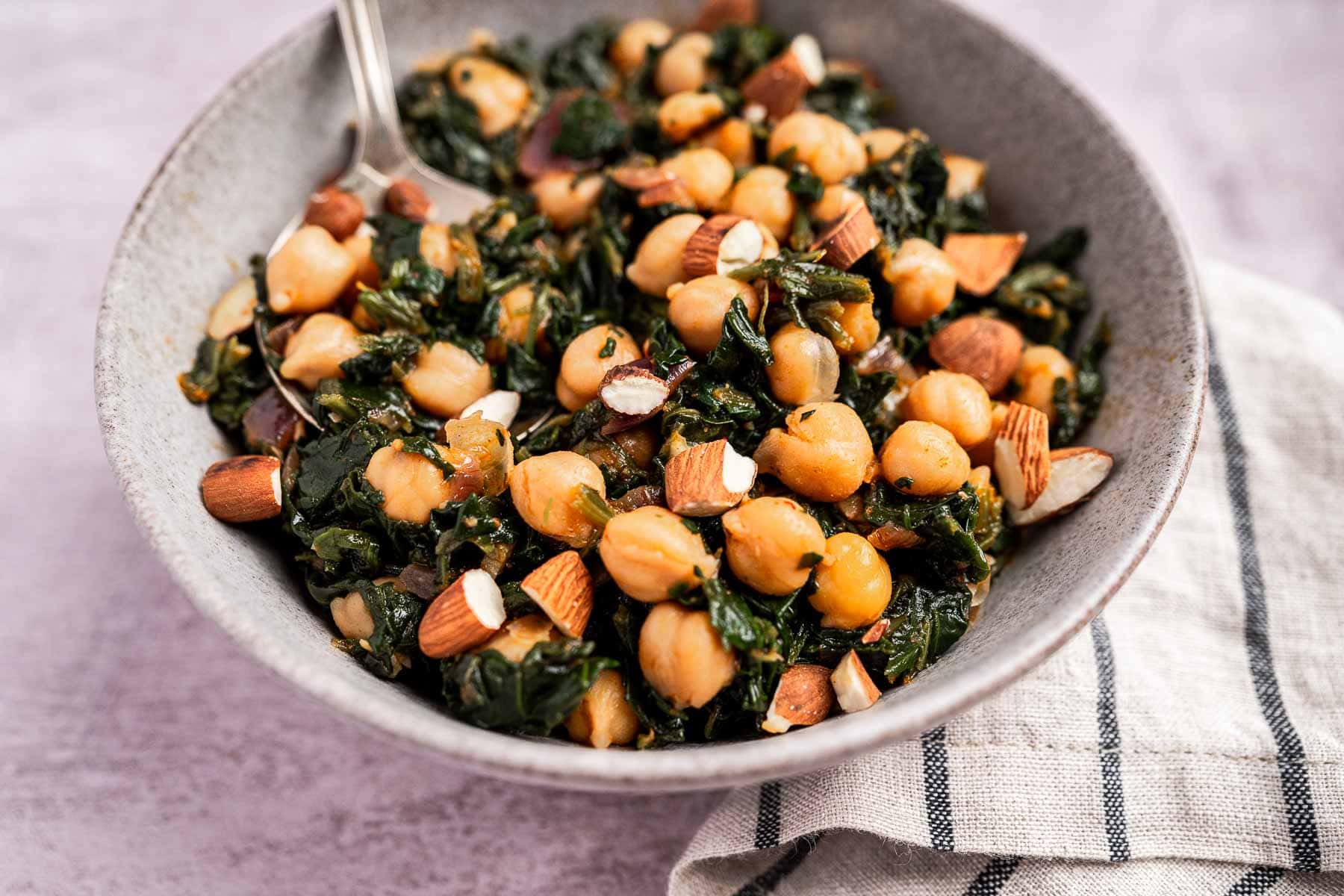 A delightful bowl of spinach and chickpeas, sprinkled with chopped almonds and accompanied by a spoon, rests invitingly on a striped cloth.