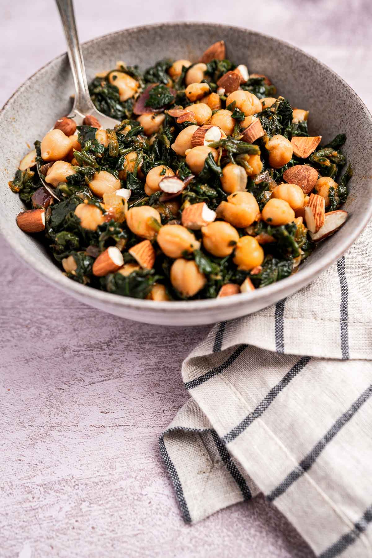 A bowl of tender chickpeas mingling with vibrant spinach and crunchy chopped almonds rests on a textured surface, accompanied by a striped cloth.