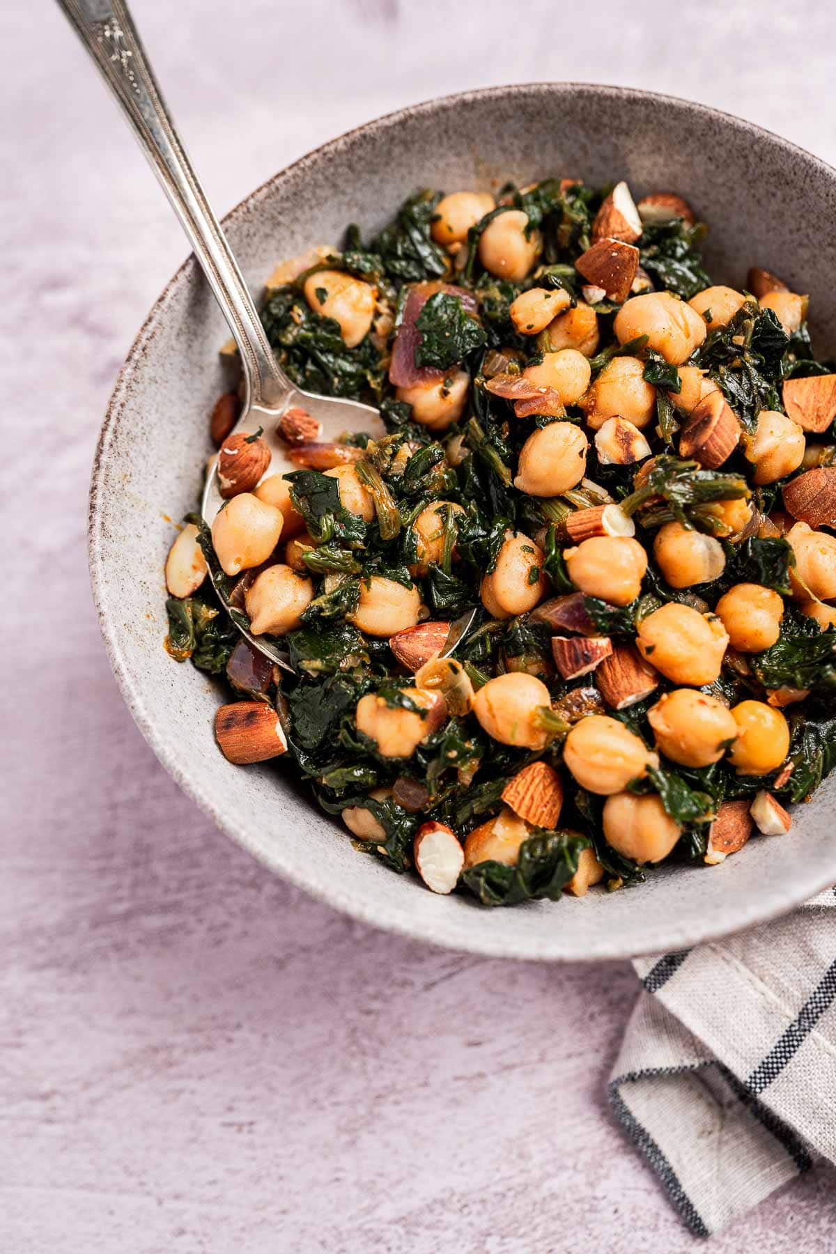 A bowl of chickpeas and vibrant spinach mixed with cooked greens and chopped almonds, a spoon resting inside.