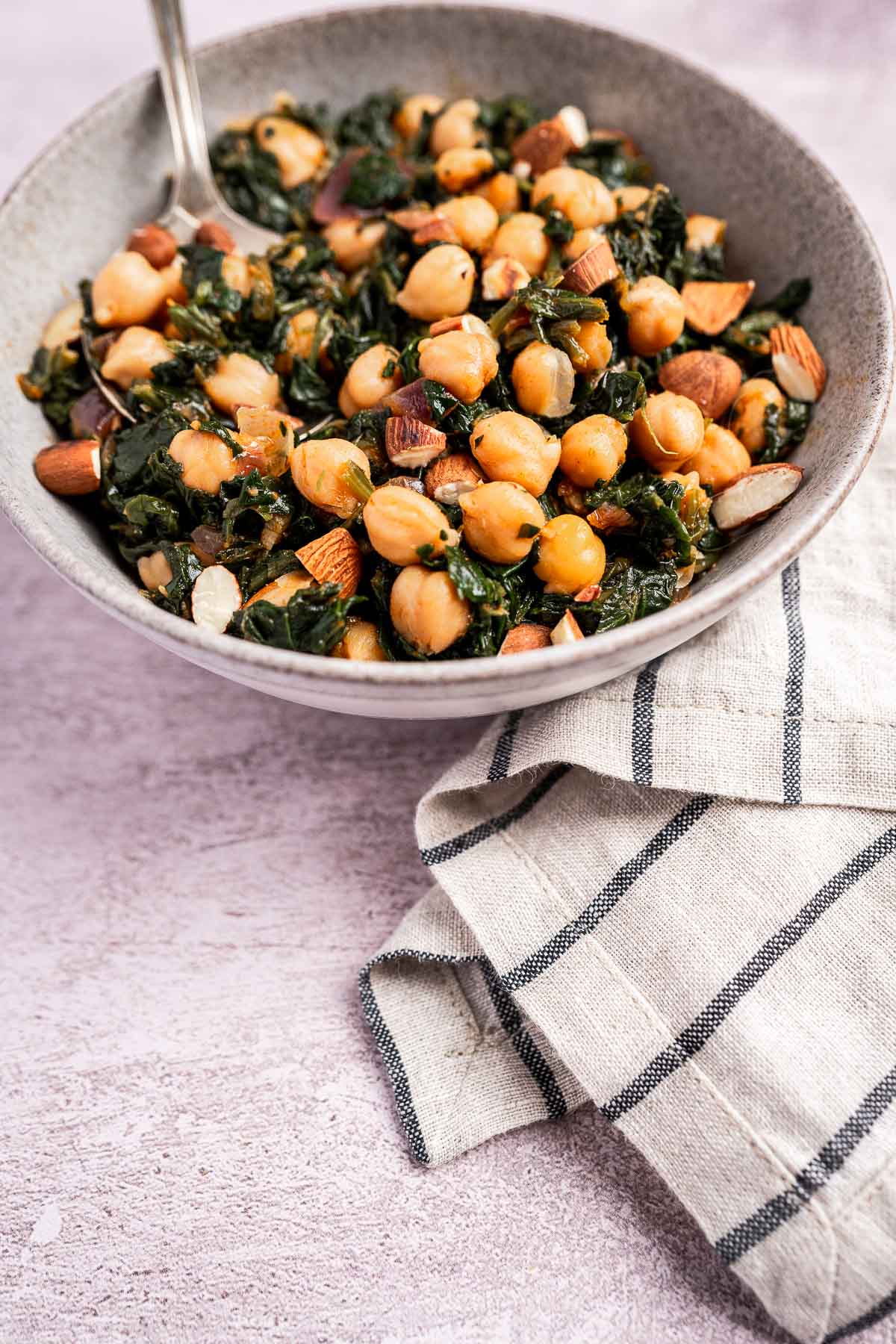 A vibrant bowl of spinach and chickpeas, sprinkled with crunchy almonds, rests elegantly on a cloth napkin with a fork nestled inside.