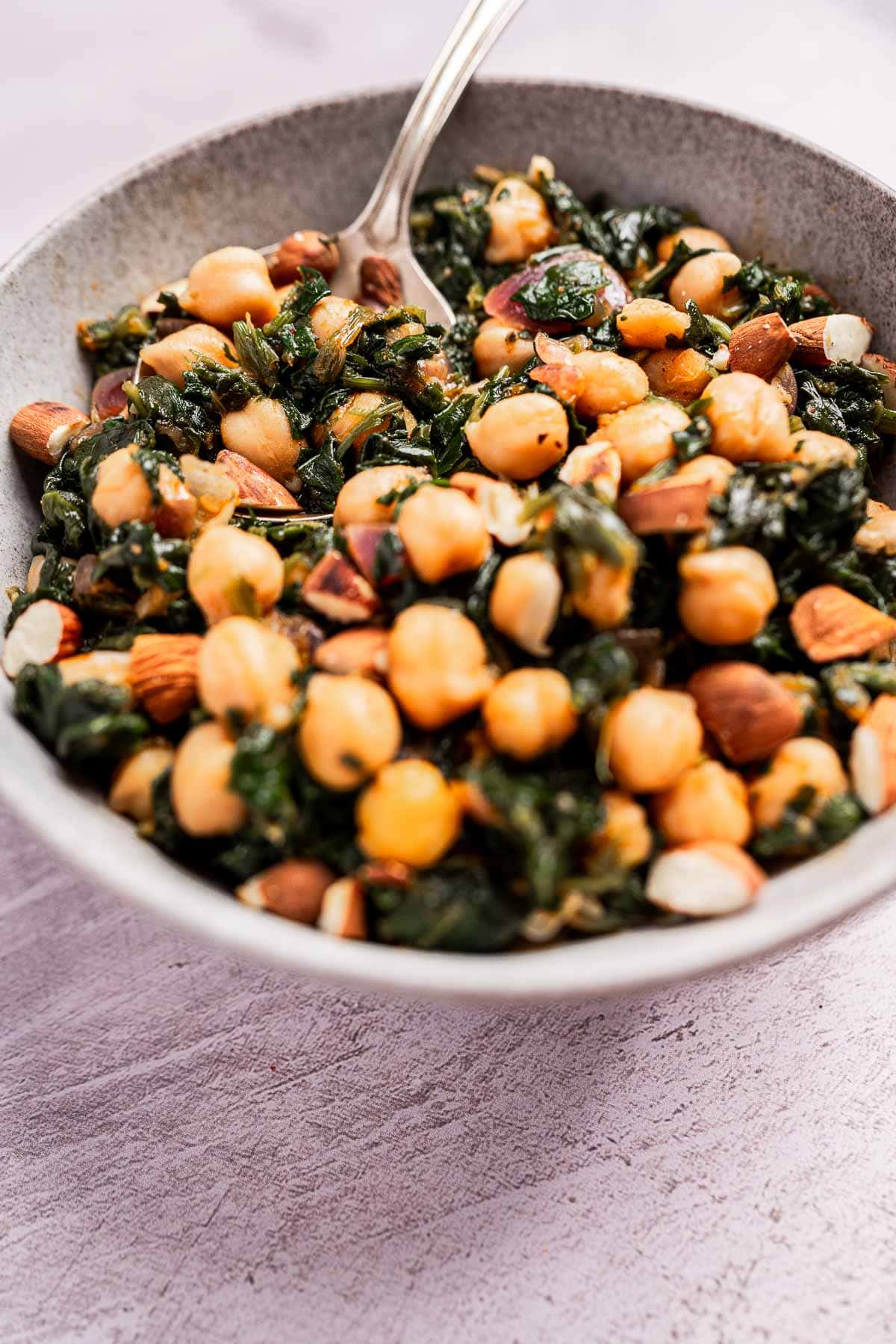 Bowl of delicious spinach and chickpea salad, garnished with almonds, and a spoon resting on a textured surface.