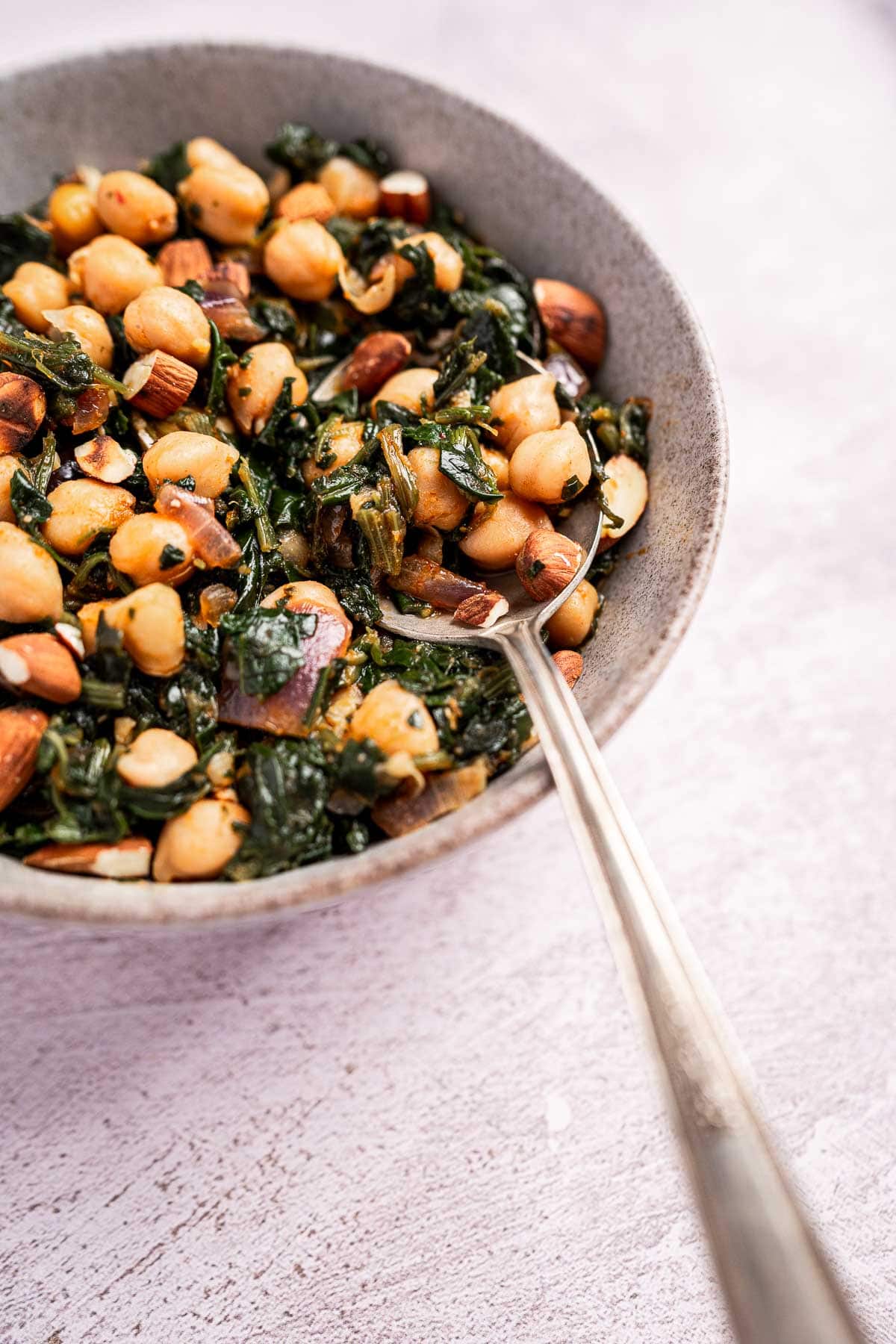 A bowl of spinach and chickpeas tossed with almonds and seasoning, served with a metal spoon on a light surface.