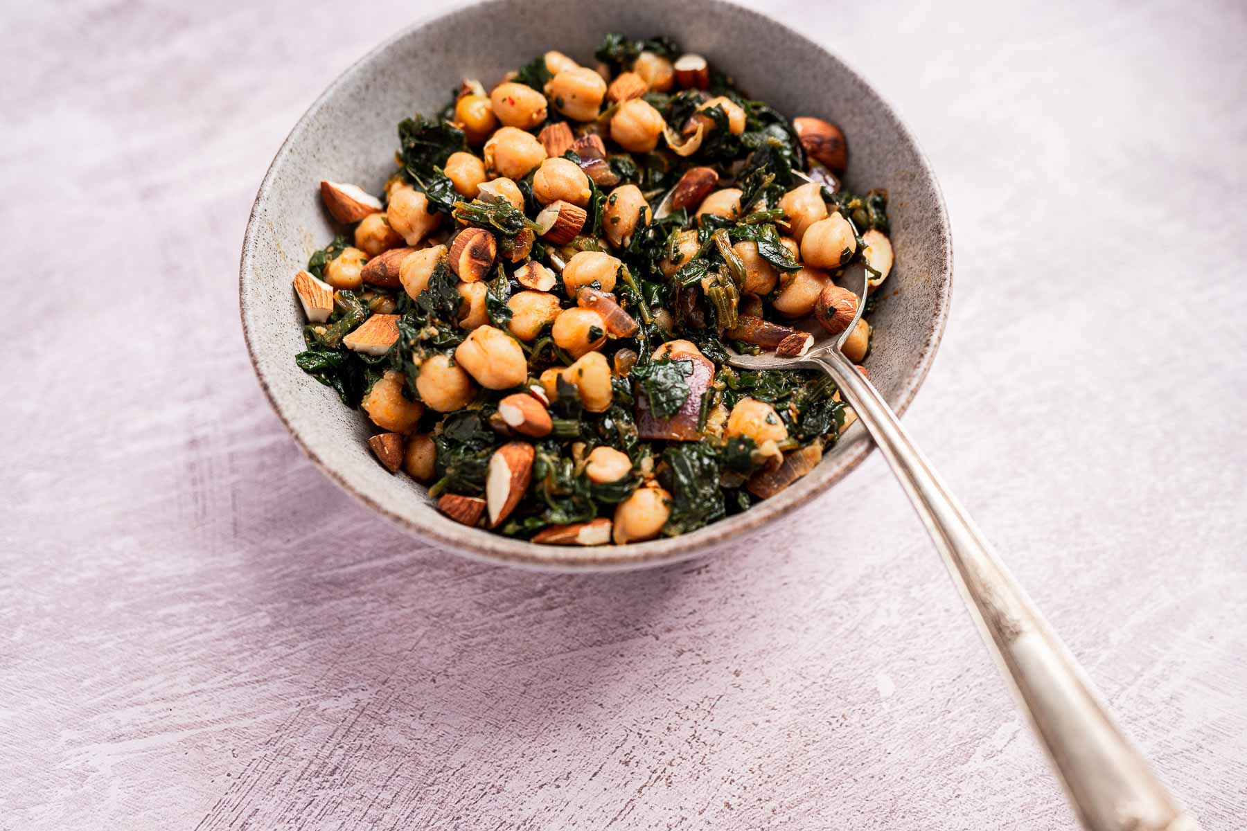 A bowl of vibrant spinach and chickpeas mingles with crunchy almonds and aromatic spices. A spoon rests beside the bowl, inviting a taste.