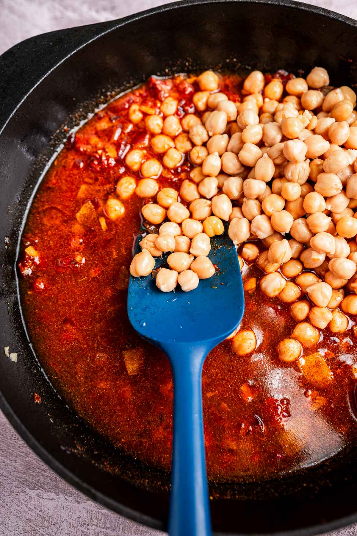 A black pan containing chickpeas and spinach in a rich red sauce, complemented by a blue spatula.