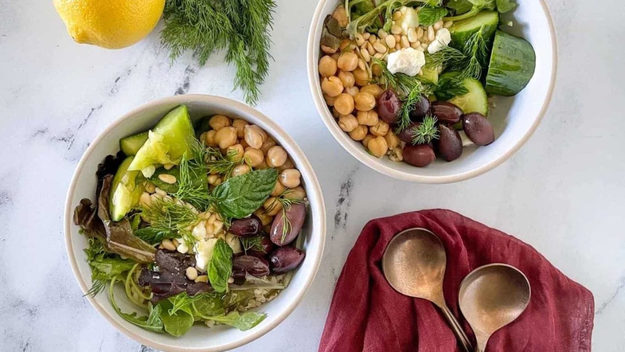 Two grain bowls are shown on a white marble counter with forks, a red linen, dill, and a lemon.
