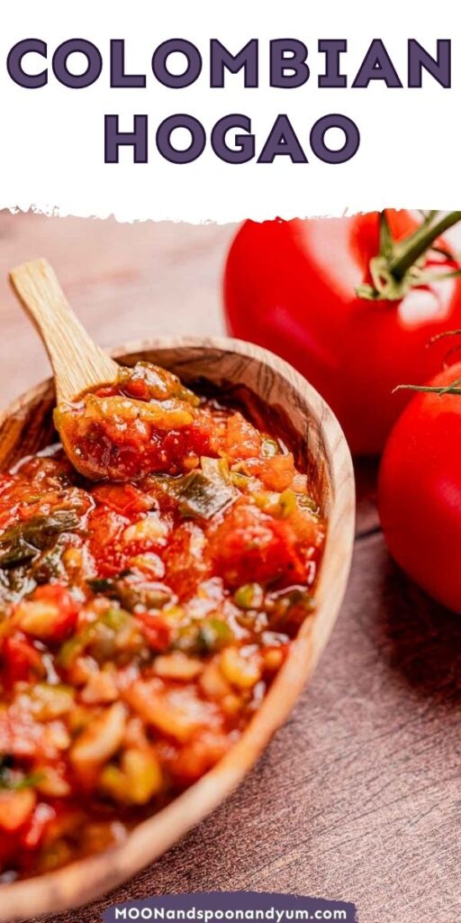A rustic wooden bowl cradles a savory Colombian hogao, a rich tomato and onion sauce, complete with a wooden spoon for serving. In the backdrop, two plump tomatoes add a touch of freshness.