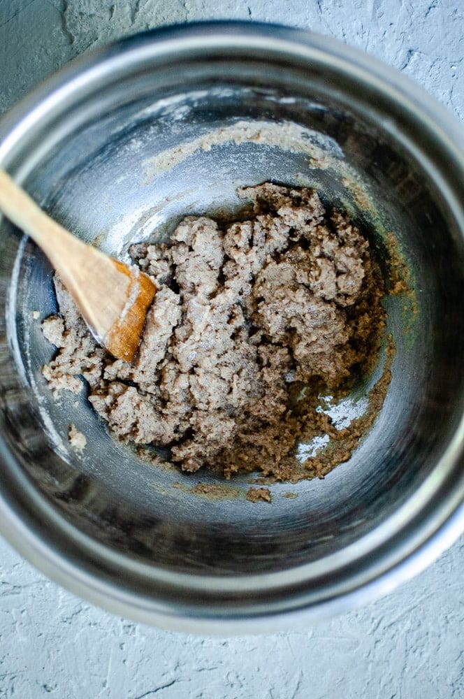 A mixing bowl with a wooden spatula stirs a thick, brown dough for sorghum flour cookies on a textured gray surface.