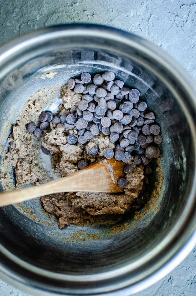 A stainless steel bowl brims with cookie dough and chocolate chips, being expertly mixed with a wooden spoon to craft delectable sorghum flour cookies.