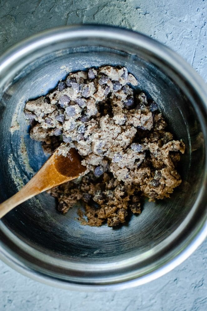 A metal bowl brimming with sorghum flour cookie dough, rich in chocolate chips, and accompanied by a trusty wooden spoon.