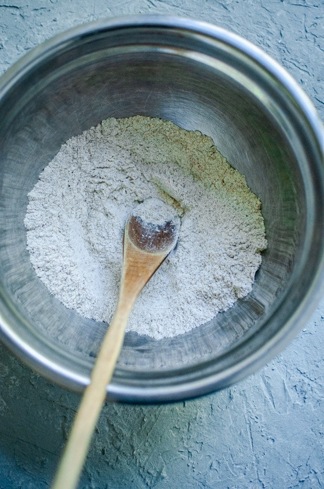 A metal mixing bowl containing flour, hinting at homemade sorghum flour cookies in the making, with a wooden spoon placed inside on a textured gray surface.
