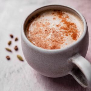 A ceramic mug filled with frothy chai latte sprinkled with cinnamon, placed on a table with a few cardamom pods nearby.