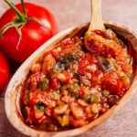 A wooden bowl filled with chunky salsa resembling traditional hogao, featuring tomatoes and green peppers, with whole tomatoes in the background.