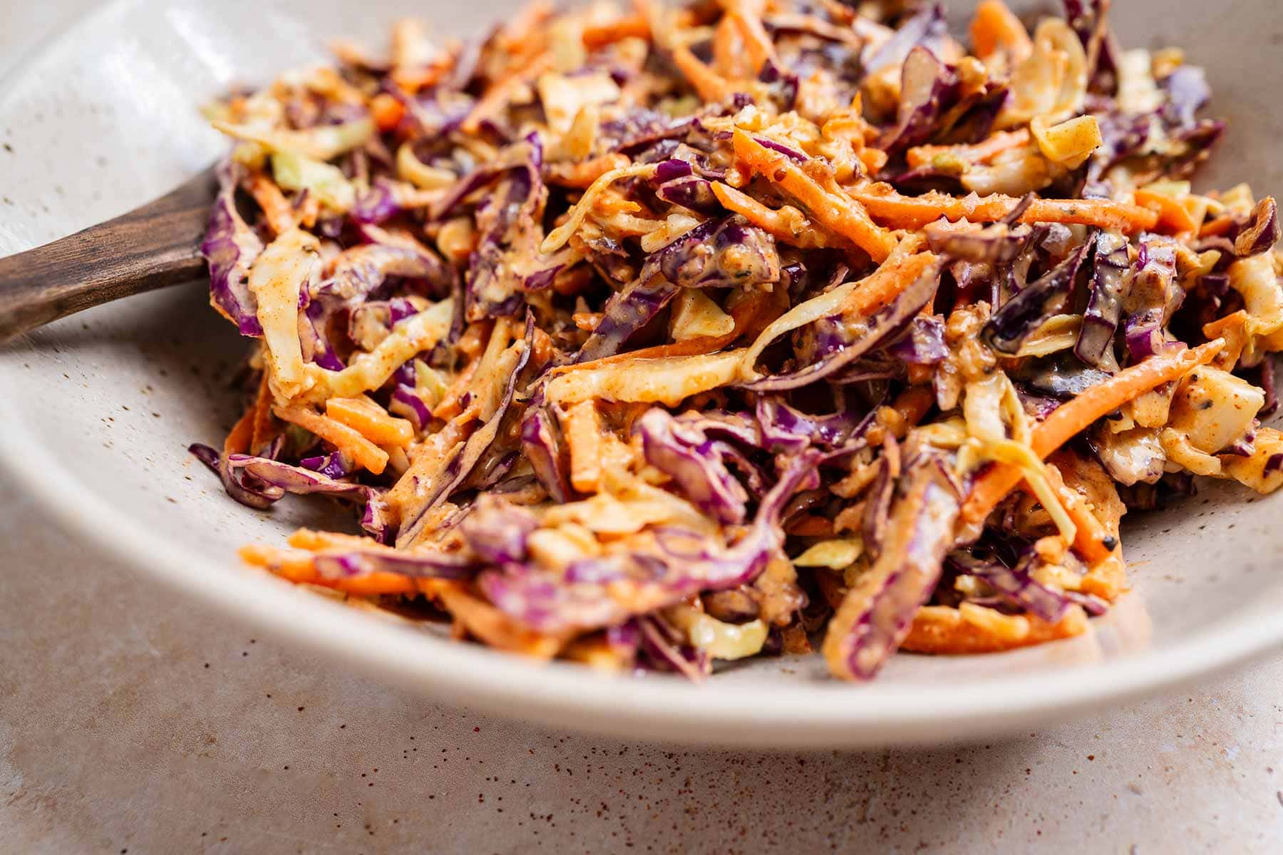 A bowl of Cajun coleslaw with shredded purple and green cabbage, carrots, and zesty dressing, served in a white dish with a wooden spoon.