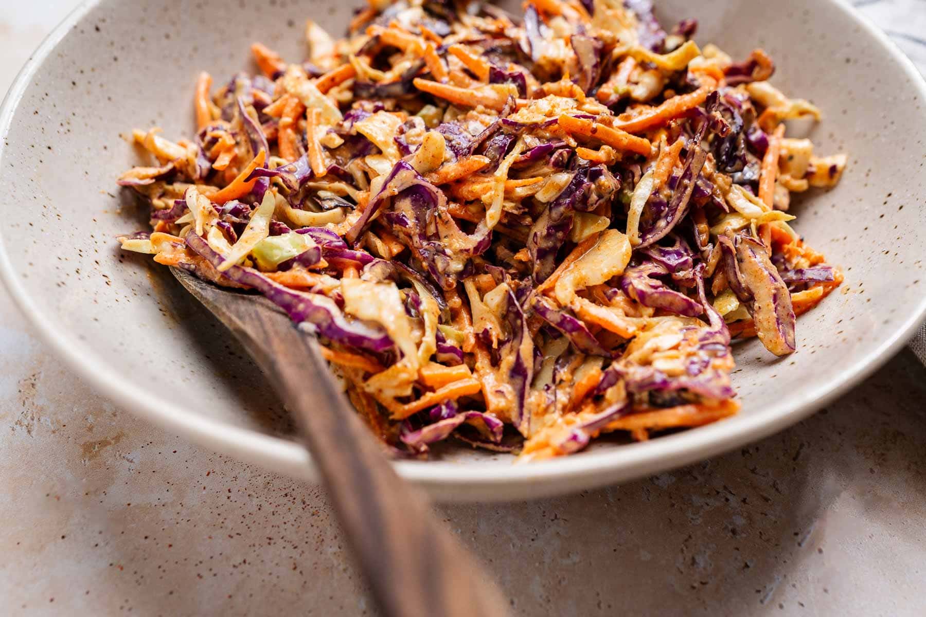 A bowl of vibrant Cajun coleslaw features shredded carrots, purple cabbage, and a creamy dressing with a hint of spice, accompanied by a wooden spoon on the side.