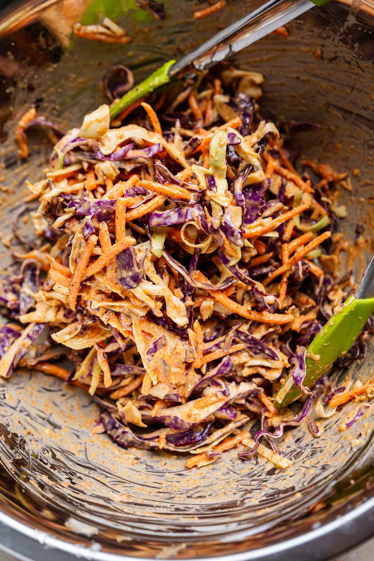 A bowl of Cajun coleslaw with shredded carrots, purple cabbage, and green cabbage, tossed in a creamy dressing with a hint of spice, metal tongs on the side.