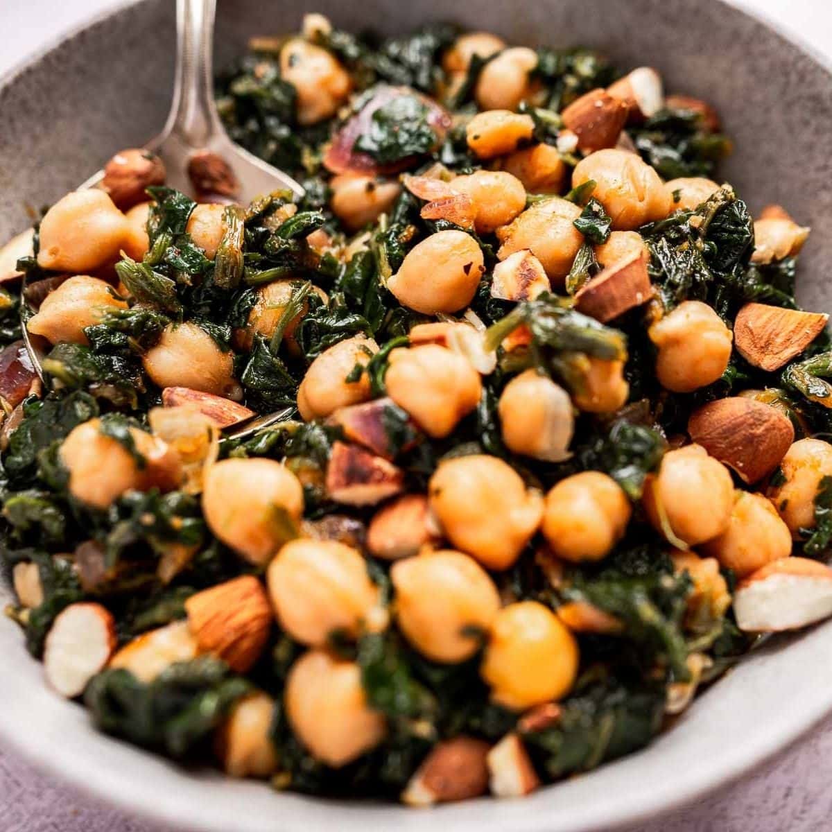 A bowl of chickpeas with spinach, almonds, and herbs, served with a silver spoon.