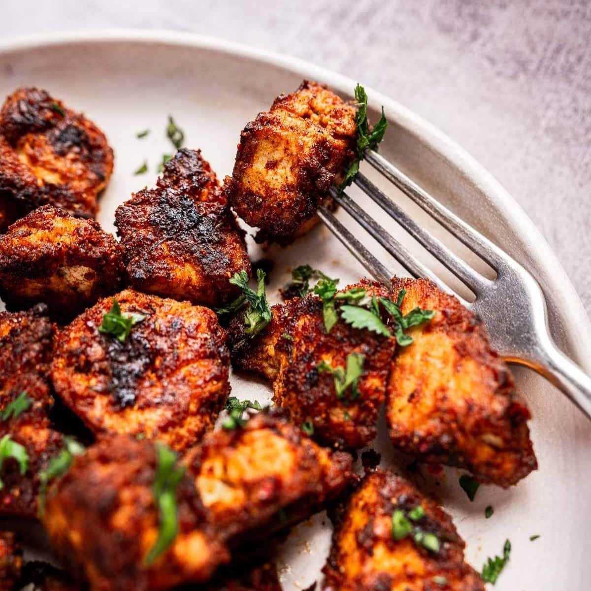 Close-up of spiced and grilled chicken pieces on a plate, garnished with chopped herbs, with a fork resting nearby.