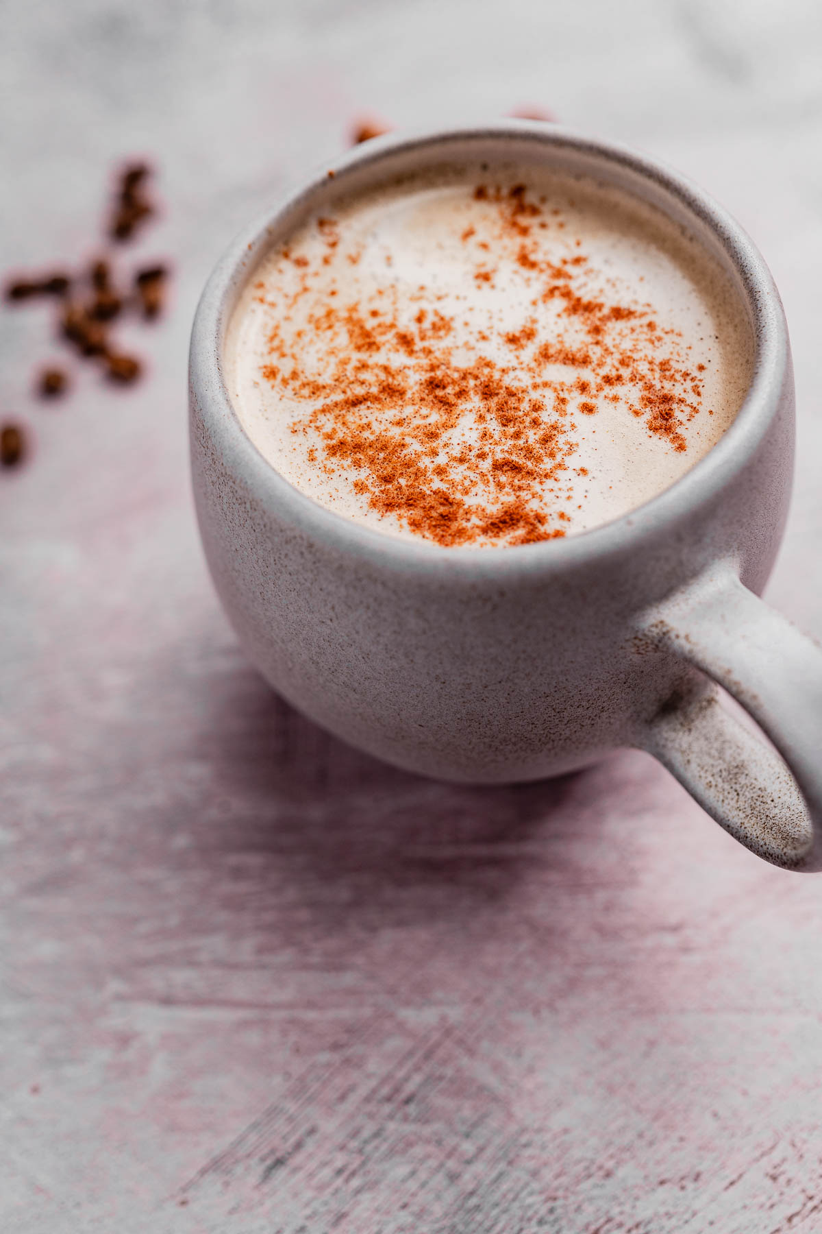 A ceramic mug filled with creamy chai coffee topped with a sprinkle of cinnamon sits on a textured surface, with scattered spice pieces in the background.
