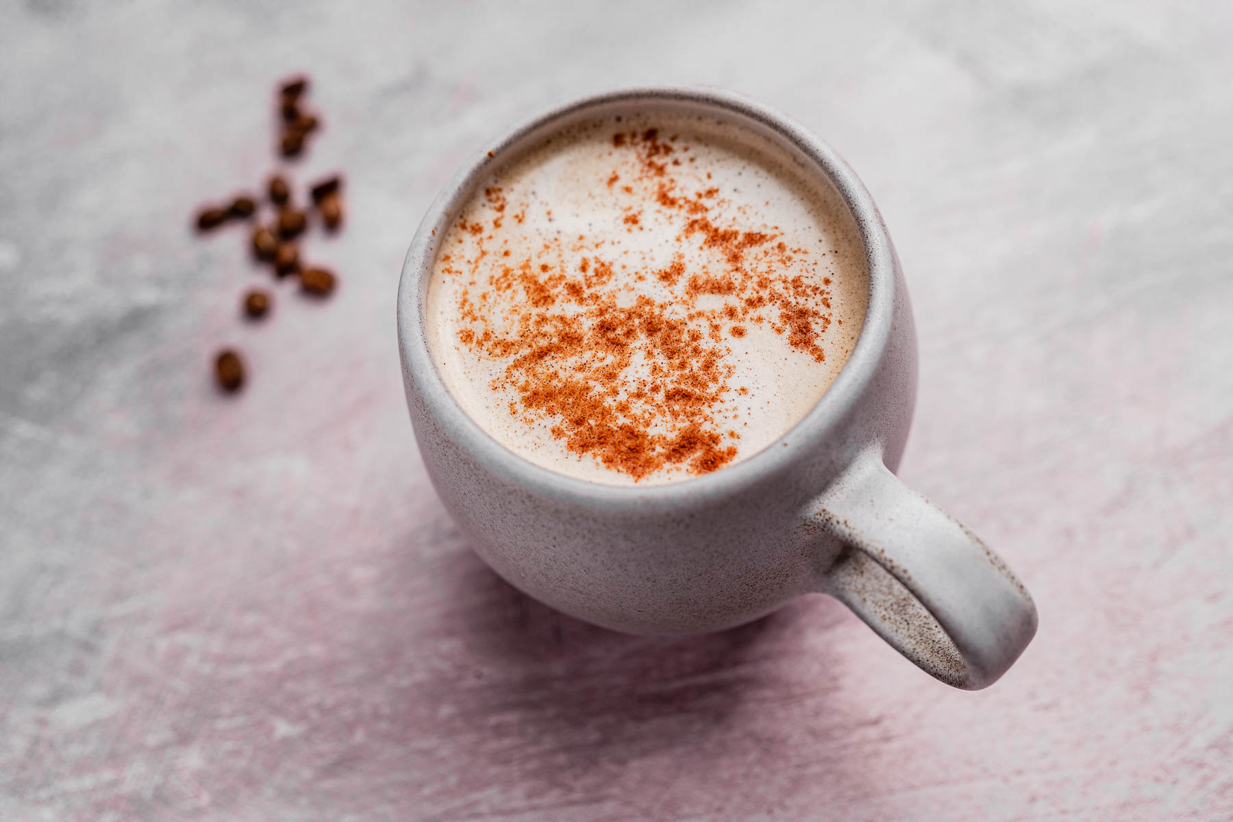 A mug of foamy chai coffee topped with sprinkled cinnamon rests on a textured surface, surrounded by a few scattered coffee beans.