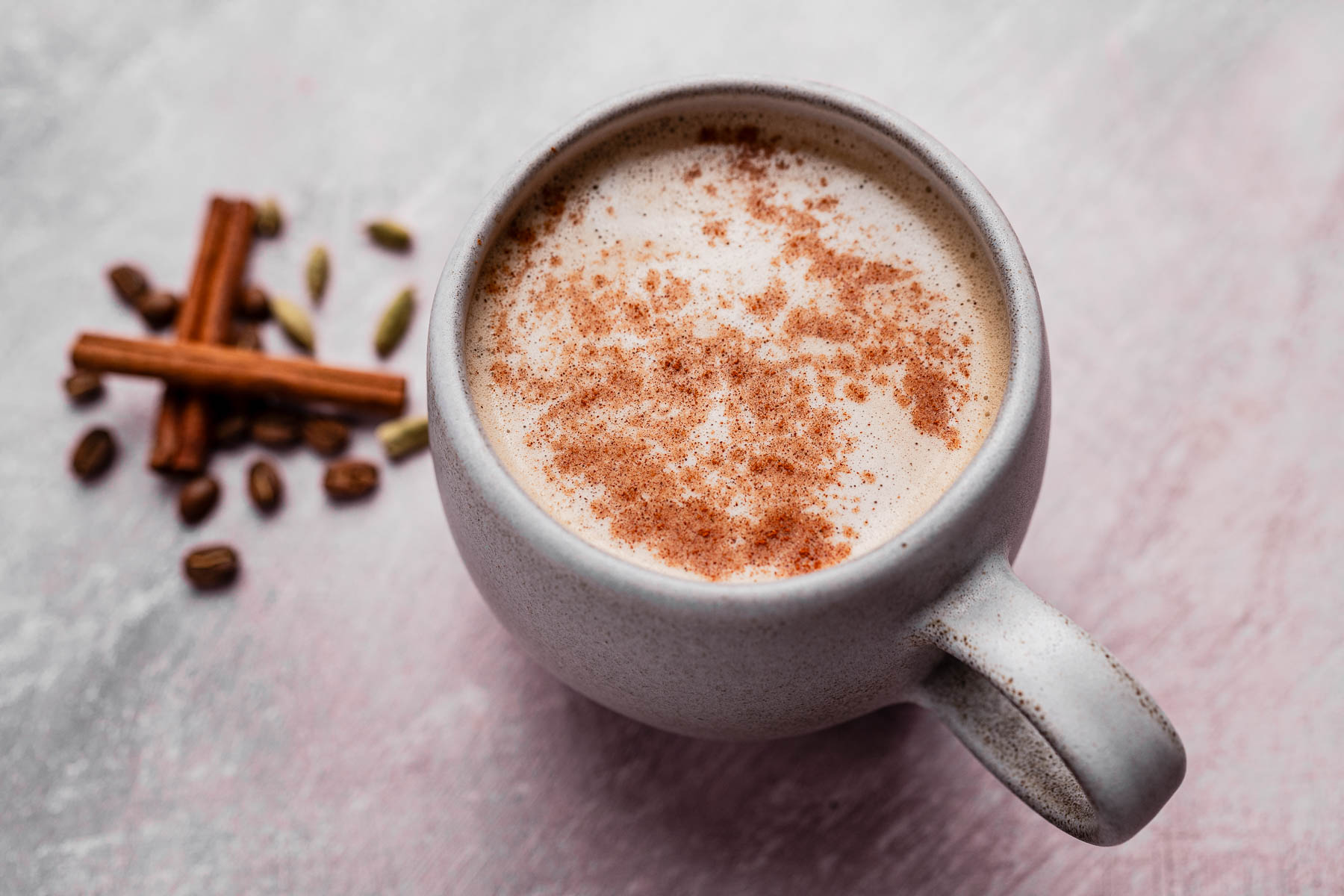 A mug of chai coffee latte topped with cinnamon sits next to cinnamon sticks, cardamom pods, and cloves on a textured surface.