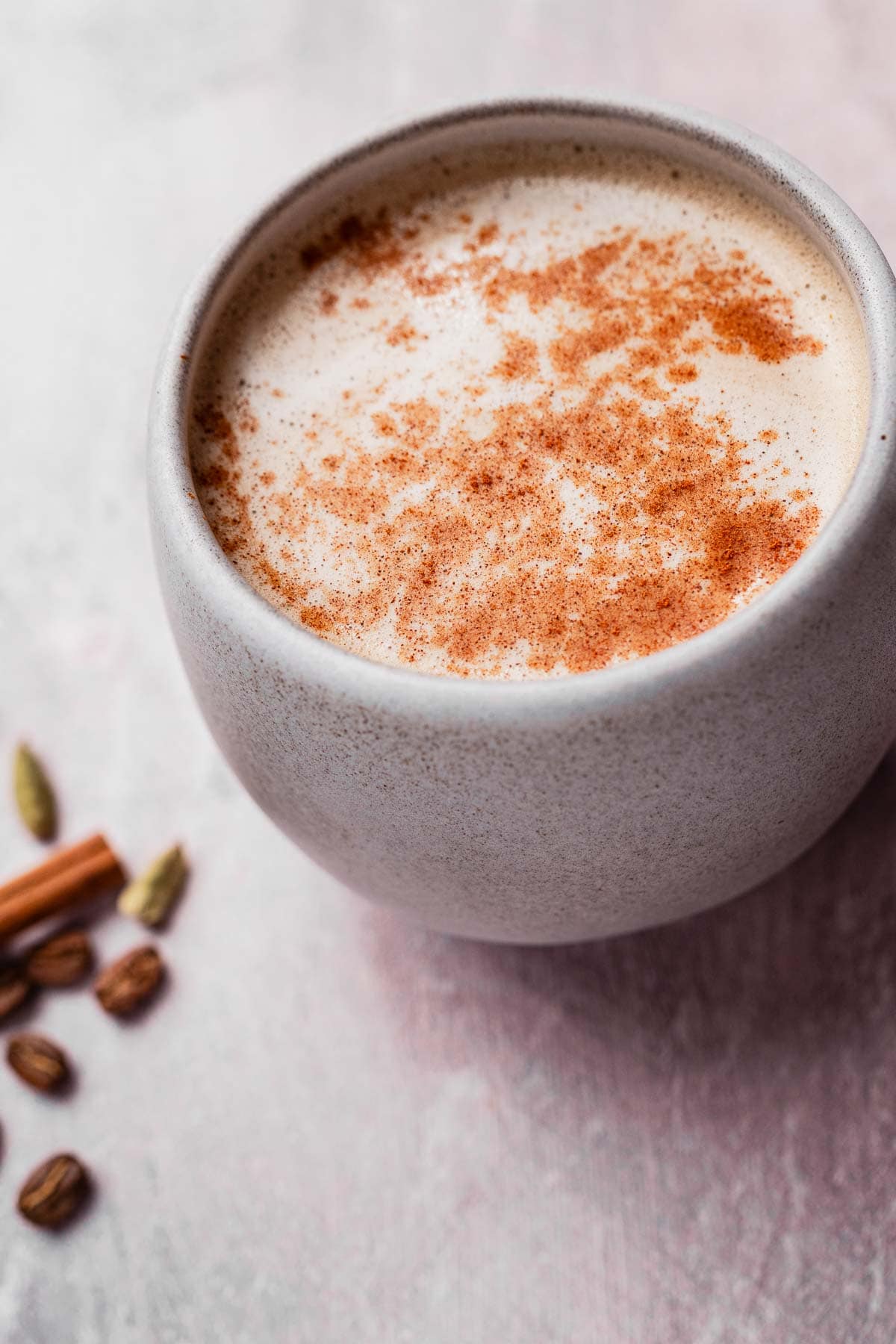 A ceramic cup filled with frothy chai coffee, topped with cinnamon powder. Nearby are coffee beans, cardamom pods, and a cinnamon stick on a light surface.
