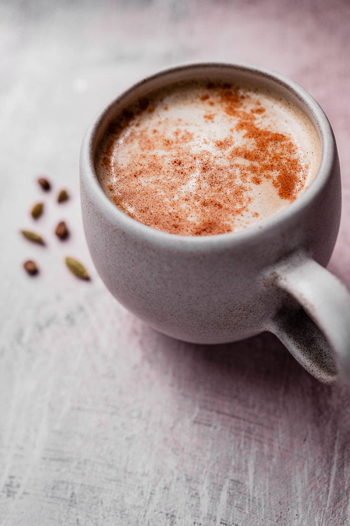 A mug filled with frothy chai coffee, topped with cinnamon, sits on a textured surface with cardamom pods scattered beside it.