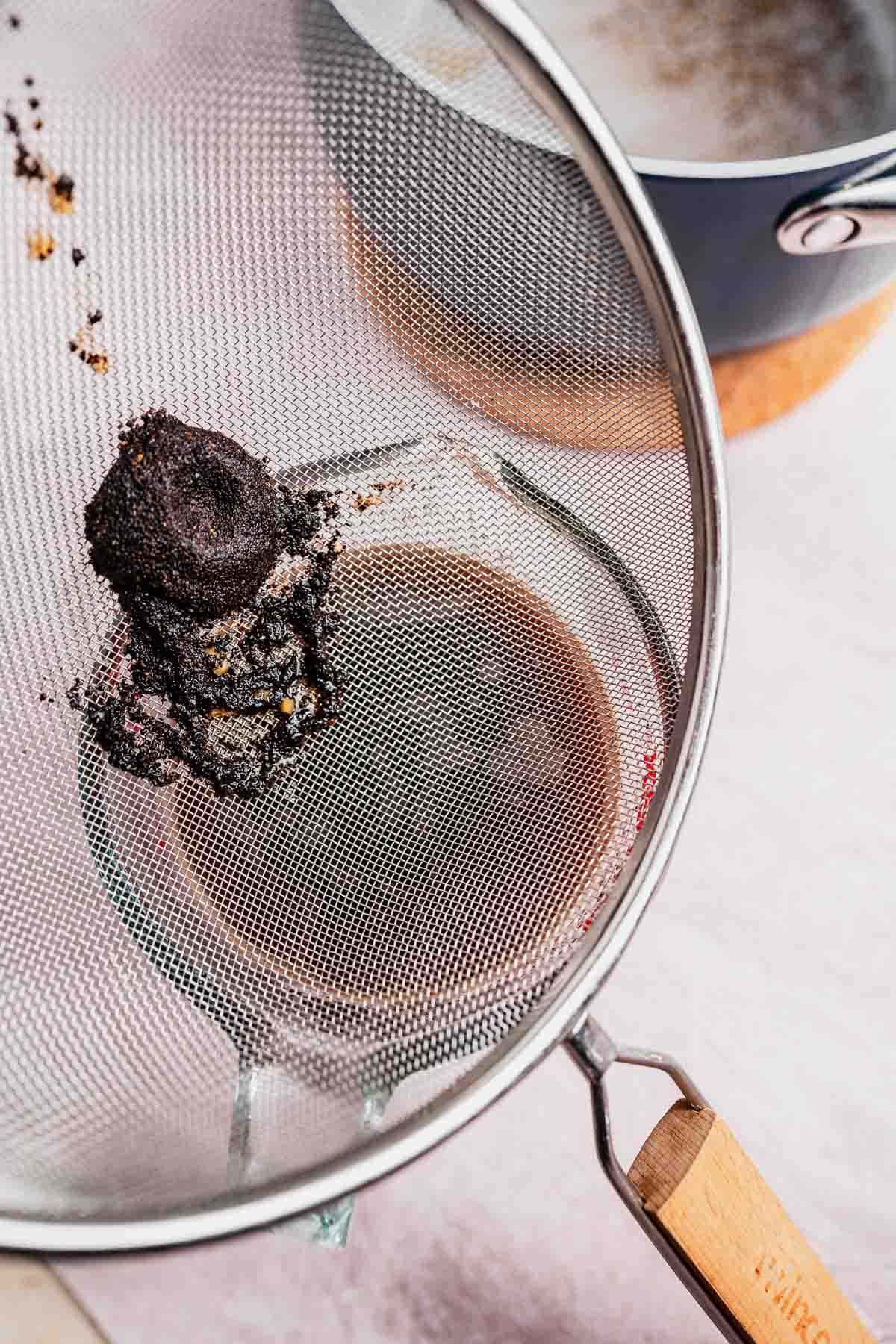 A fine mesh strainer with used tea leaves rests over a glass jar containing chai, beside a saucepan on a countertop.