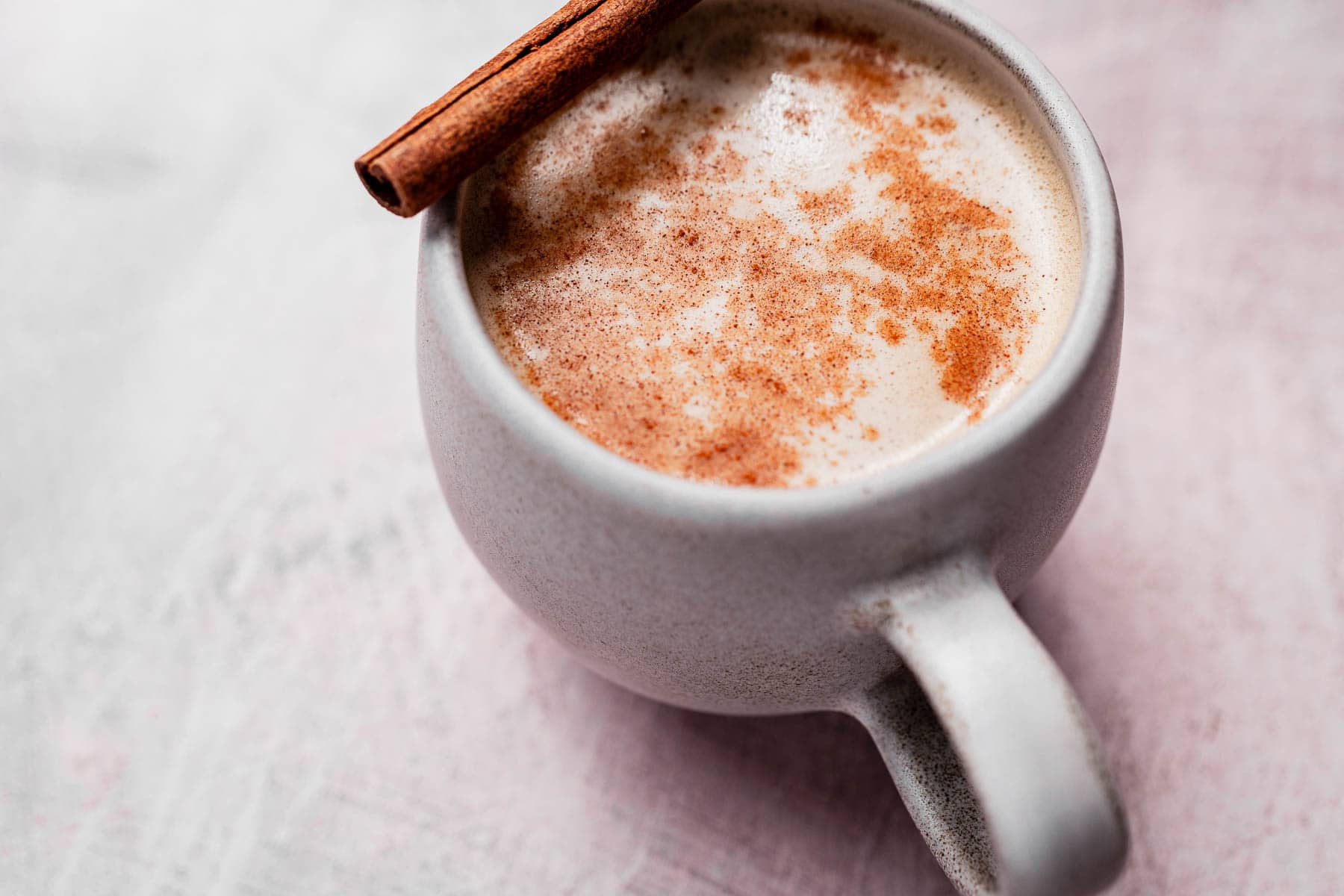 A frothy cup of chai coffee with cinnamon sprinkled on top, garnished with a cinnamon stick, rests invitingly on a light-colored surface.