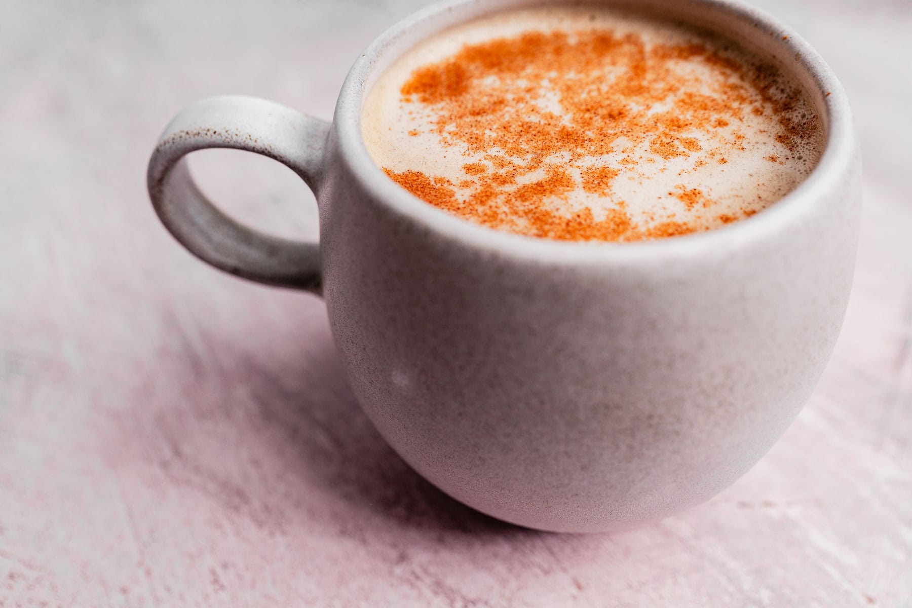 A ceramic cup filled with frothy chai coffee, topped with a sprinkle of cinnamon, sits on a textured pink surface.