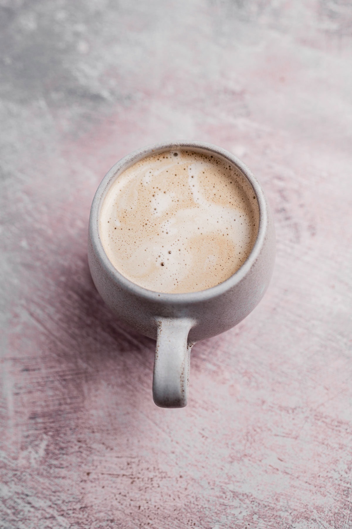 A cup of frothy chai coffee with a creamy swirl in a gray ceramic mug, placed on a textured pink and gray surface.