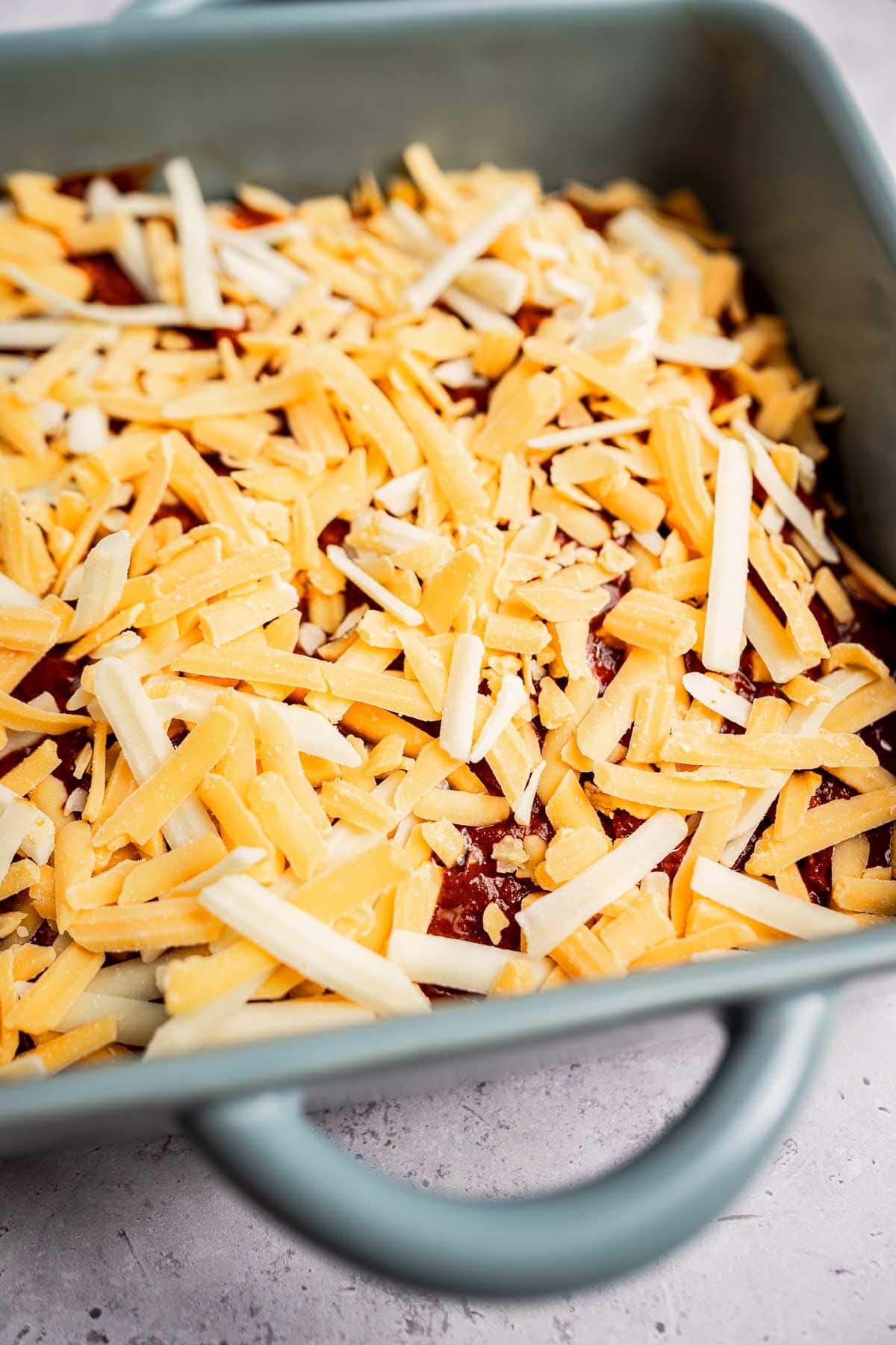 A blue baking dish brimming with shredded cheese sits on a reddish-brown base, ready to transform into a delectable cheese enchiladas casserole.
